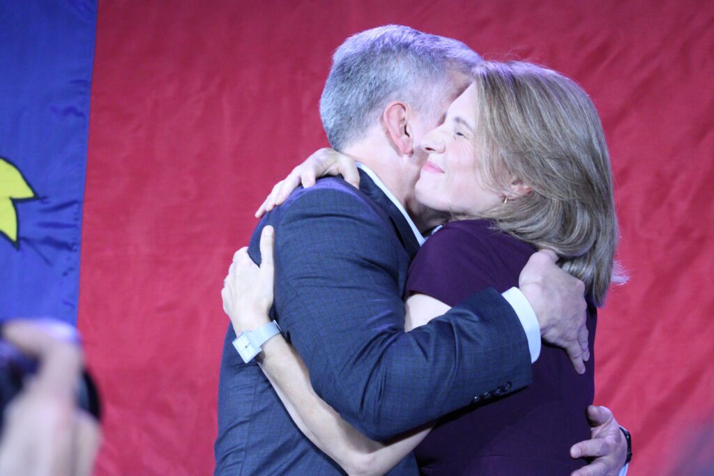 Josh Stein hugs wife with both arms in front of North Carolina flag