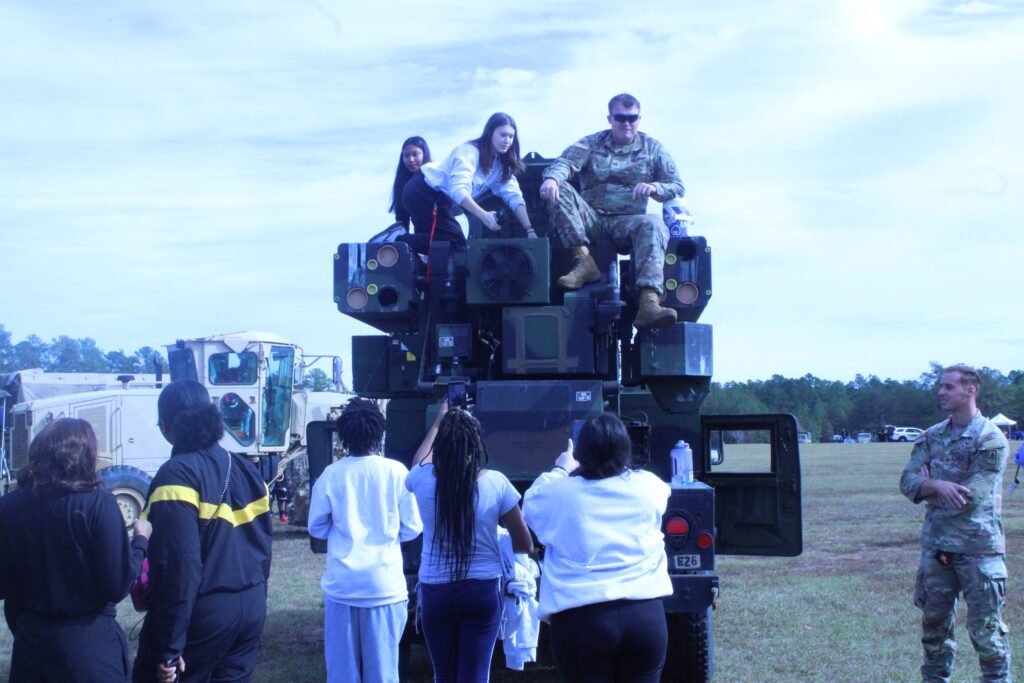 students sitting on a black truck with a soldier in camouflage